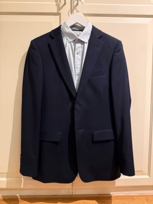 Dressmann kostym + Jack & Jones skjorta - Säljer en stilren marinblå kostym från Dressmann tillsammans med en ljusblå skjorta från Jack & Jones premiumsortiment. Passar perfekt till bal, student eller fest med mera. Skjorta: medium, slim fit. Kavaj: slim fit, long, storlek 46. Kostymbyxor: 31-32.  Priset kan diskuteras vid snabb affär.