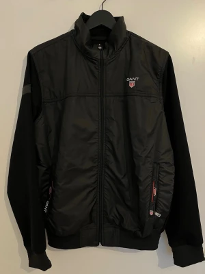 Svart Gant vindjacka med logga - Stilren svart vindjacka från Gant med hög krage och dragkedja framtill. Jackan har två fickor med dragkedja och röda detaljer, samt Gant-logga på bröstet. Framsidan är i glansigt material medan ärmar och rygg är i mjukare tyg. Perfekt för en sportig och clean look.