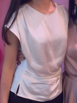 Ljusbeige silk omlottopp med knyt - Säljer en ljusbeige omlottopp i mjukt, glansigt material. Toppen har korta ärmar, rund halsringning och en snygg knytdetalj i sidan som ger en cool twist. Perfekt att styla med jeans eller kjol för en trendig look.