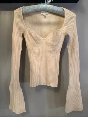Beige ribbad långärmad topp - Trendig beige ribbad topp med lång ärm och hjärtformad ringning. Perfekt för att styla med jeans eller kjol för en snygg look.