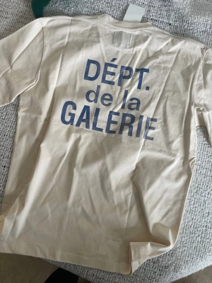 Beige t-shirt från Gallery Dept. - Snygg beige t-shirt från Gallery Dept. med blå text 'DÉPT. de la GALERIE' tryckt både fram och bak. Klassisk passform med rund hals och korta ärmar. Tillverkad i mjuk bomull som känns skön mot huden. Perfekt för en avslappnad och trendig look.
