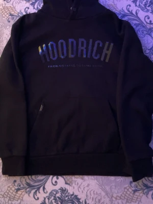 Svart hoodie från Hoodrich - Snygg svart hoodie från Hoodrich med stor logga i metallic på bröstet och texten 'FROM NOTHING TO SOMETHING' under. Klassisk känguruficka framtill och ribbade muddar vid ärmslut och nederkant. Perfekt för dig som gillar streetwear.