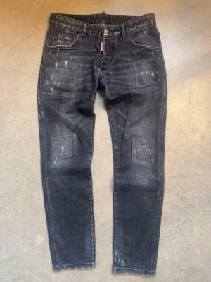 Dsquared2 jeans - Säljer ett par svarta jeans från Dsquared2 med coola slitningar och tvättad look. Jeansen har klassisk femficksmodell, knappgylf och en röd Dsquared2-logga vid fickan. Perfekta för dig som gillar streetwear och en avslappnad stil.