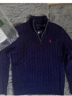 Mörkblå kabelstickad quarter zip polo - Säljer en mörkblå kabelstickad tröja från Polo Ralph Lauren med halv dragkedja och röd broderad logga på bröstet. Tröjan har hög krage, ribbade muddar och är tillverkad i mjukt material. Perfekt för dig som gillar klassisk och stilren look.