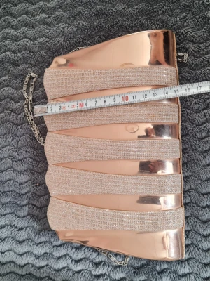 Glittrig roséguld kuvertväska - Snygg kuvertväska i roséguld med glittriga band och metallisk finish. Väskan har en kedjerem som gör att den kan bäras både i handen och över axeln. Perfekt accessoar för att lyfta din outfit med lite extra bling.