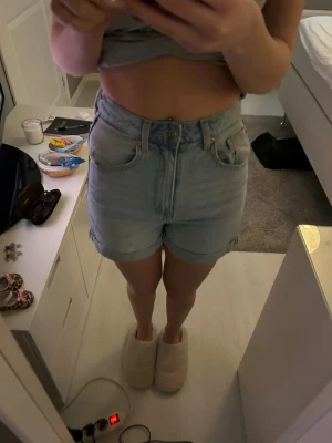 Ljusblå högmidjade jeansshorts - Säljer ett par ljusblå denimshorts med hög midja och klassisk femficksdesign. Shortsen har uppvikta benslut och stängs med knapp och dragkedja. Perfekta till sommaren och lätta att matcha med olika toppar.