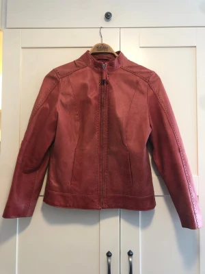 Röd skinnjacka med ståkrage - Snygg röd skinnjacka med ståkrage och dragkedja framtill. Jackan har dekorativa perforerade detaljer längs sömmarna och en stilren, figurnära passform. Perfekt för dig som vill sticka ut med en unik jacka i skinn.