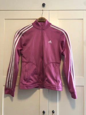 Lila Adidas träningsjacka - Snygg lila träningsjacka från Adidas med klassiska vita ränder längs ärmarna och vit Adidas-logga på bröstet. Jackan har hel dragkedja, hög krage och två fickor framtill. Perfekt för träning eller chill dagar.