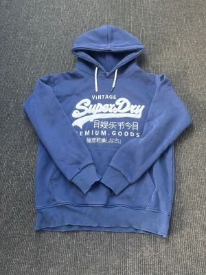 Blå Superdry hoodie  - Snygg blå hoodie från Superdry med stort vitt tryck på bröstet och japanska tecken. Klassisk känguruficka framtill, ribbade muddar och vit dragsko i huvan. Perfekt för en avslappnad och trendig stil. Fint skick, storlek L men passar M