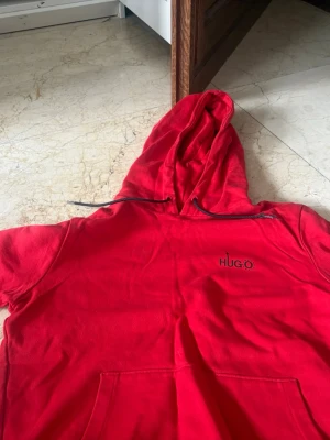 Röd hoodie från HUGO Berlin - Snygg röd hoodie från HUGO Berlin med svart logga och stjärnmönster på ryggen. Hoodien har huva med svarta snören och en stor magficka framtill. Tillverkad i mjukt bomullstyg, perfekt för en avslappnad streetwear-look.