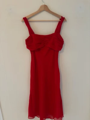 Röd långklänning med breda axelband - Snygg röd mididress med breda axelband och volangdetaljer upptill. Klänningen har en figurnära passform och är tillverkad i ett lätt, flytande material som ger ett elegant intryck. Perfekt för dig som vill sticka ut med en klassisk och stilren look.