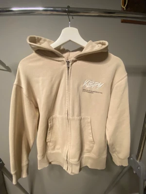 Beige zip hoodie från KBPY - Snygg beige hoodie med dragkedja från KBPY. Hoodien har stor huva, fickor framtill och tryck i vitt både på bröstet och ryggen. Tillverkad i mjukt bomullsmaterial och har ribbade muddar vid ärmslut och nederkant. Perfekt för en avslappnad streetwear-look.
