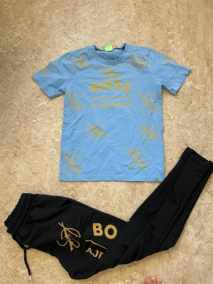 Blått och svart set med BOSS-tryck - Säljer ett tvådelat set med en ljusblå t-shirt och svarta byxor. Båda delarna har gula tryck med texten BOSS och AJBKNB samt grafiska detaljer. T-shirten har rund hals och korta ärmar, byxorna har resår i midjan och dragsko. Materialet ser ut att vara bomull.