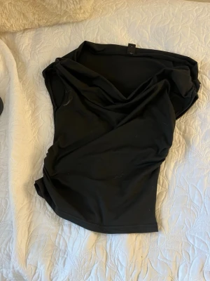 Svart ärmlös topp med drapering - Snygg svart topp med draperad v-ringning och ärmlös design. Perfekt att styla med jeans eller kjol för en trendig look. Mjuk och stretchig känsla i materialet.