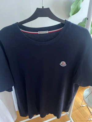 Moncler T-shirt  - Snygg marinblå t-shirt från Moncler med klassisk rund halsringning och diskret Moncler-logga på bröstet. Tillverkad i mjuk bomull som känns skön mot huden. Perfekt basplagg med stilren design och korta ärmar.