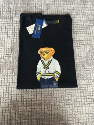 Svart Polo Bear t-shirt från Ralph Lauren - Snygg svart t-shirt från Ralph Lauren med den ikoniska Polo Bear-figuren tryckt på bröstet. T-shirten har rund hals, korta ärmar och är gjord i mjuk bomull. Perfekt för dig som gillar klassisk men lekfull stil.