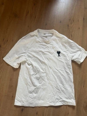 Vit t-shirt från Ami Paris - Vit t-shirt från Ami Paris med svart hjärta logga broderad på bröstet. Klassisk passform och rund halsringning. Tillverkad i bomull och har korta ärmar. Perfekt basplagg med en stilren detalj framtill. Pris kan diskuteras