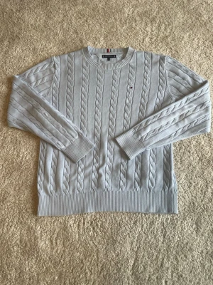 Ljusgrå kabelstickad tröja Tommy Hilfiger - Snygg ljusgrå kabelstickad tröja från Tommy Hilfiger med rund halsringning och diskret logga broderad på bröstet. Tröjan har ribbade muddar vid ärmslut och nederkant. Perfekt för dig som gillar klassisk och stilren design.