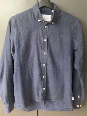 Blå manchesterskjorta från Les Deux - Stilren blå manchesterskjorta från Les Deux med klassisk krage och vita knappar. Skjortan har lång ärm och diskret broderad logga på bröstet. Perfekt för dig som gillar en avslappnad men ändå snygg look.