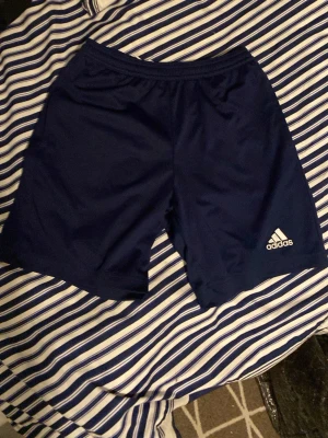 Mörkblå träningsshorts från Adidas - Snygga mörkblå träningsshorts från Adidas med vit logga på vänster ben. Tillverkade i lätt och snabbtorkande polyester, perfekta för sport eller gym. Klassisk rak passform och elastisk midja för skön komfort.