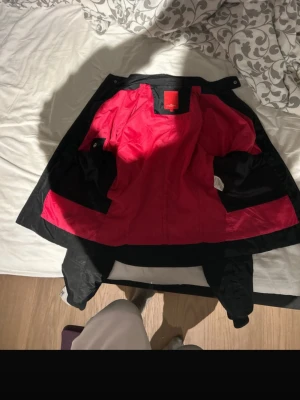 Svart vindjacka från J.Lindeberg - Stilren svart vindjacka från J.Lindeberg med röd insida. Jackan har en klassisk passform och är tillverkad i lätt och slitstarkt material, perfekt för blåsiga dagar. Märkeslogga på insidan och praktiska fickor. Snygg och funktionell design.