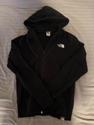 Svart zip hoodie från The North Face - Snygg svart zip hoodie från The North Face med dragkedja framtill och vit logga på bröstet och ryggen. Hoodien har justerbar huva med snören, ribbade muddar och är tillverkad i mjukt bomullsmaterial. Perfekt för en avslappnad och cool stil.
