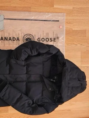 Svart dunjacka från Canada Goose - Snygg svart dunjacka från Canada Goose med huva och quiltad design. Jackan har dragkedja och tryckknappar framtill samt justerbar huva. Perfekt för kalla vinterdagar och riktigt varm tack vare dunfyllningen.
