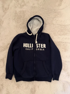Marinblå hoodie från Hollister - Mysig marinblå hoodie från Hollister med dragkedja och vit text 'Hollister California' på bröstet. Utrustad med stor huva, vita dragsnören och två fickor framtill. Insidan är fodrad i mjukt, ljust ull i storlek M-L