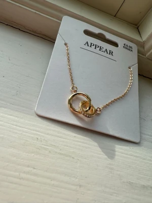 Guldigt halsband med ringar från Appear - Snyggt halsband i guldfärg från Appear med två sammanlänkade ringar, där den ena ringen är dekorerad med små gnistrande stenar. Kedjan är tunn och elegant, perfekt för att lyfta vilken outfit som helst.