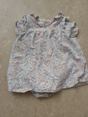 Klänning - Söt babyklänning i ljusa färger med blommigt mönster i rosa, blått och vitt. Klänningen har volangärmar och en lös, bekväm passform. Tillverkad i mjukt bomullstyg, perfekt för små barn.