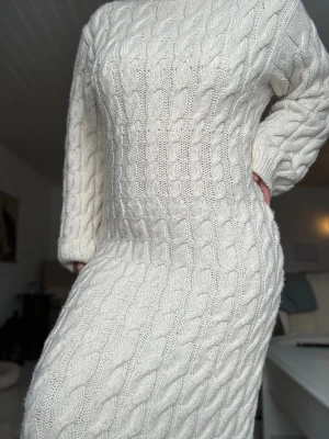 Vit lång stickad klänning från H&M - Mysig vit lång stickad klänning med kabelstickat mönster och långa ärmar från H&M. Perfekt för kyligare dagar och ger en avslappnad men ändå stilren look. Klänningen har en kroppsnära passform och är tillverkad i ett mjukt material.