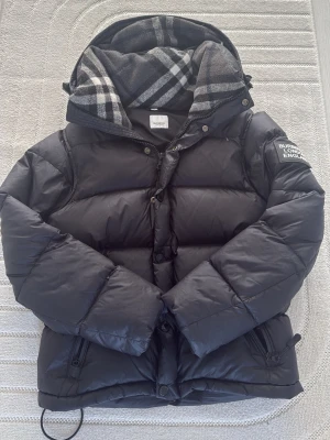 Svart dunjacka från Burberry - Snygg svart dunjacka från Burberry med klassiskt rutigt foder i huvan. Jackan har dragkedja och knappar framtill, två fickor med dragkedja och en patch med Burberry-logga på ärmen. Perfekt för kalla vinterdagar och riktigt stilren look.