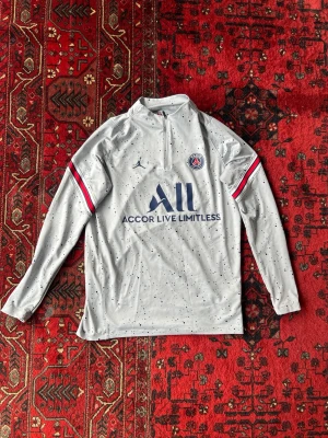 Grå PSG träningströja från Jordan - Snygg grå långärmad träningströja från PSG x Jordan med svartprickigt mönster, halv dragkedja och klubbmärke på bröstet. Röd, vit och blå detalj på ärmarna och trycket 'Accor Live Limitless' framtill. Perfekt för fotbollsträning eller chill.
