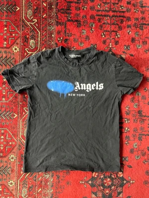 Svart Palm Angels t-shirt med tryck - Säljer en svart t-shirt från Palm Angels med ikoniskt tryck framtill där det står 'Angels New York' och blå spray paint-detalj. T-shirten har rund hals och korta ärmar. Perfekt för dig som gillar streetwear och vill sticka ut.