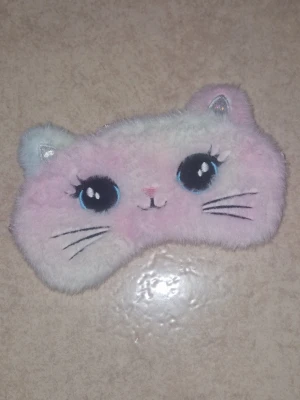 Sovmask - Supersöt sovmask i fluffigt material med kattansikte, broderade morrhår och öron. Framsidan är ljusrosa och vit med stora ögon, baksidan i mjuk satin och elastiskt band för bekväm passform. Perfekt för dig som vill ha en gullig accessoar till sovstunden.