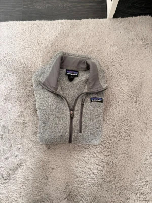 Patagonia grå väst - Säljer en grå fleecejacka från Patagonia med halv dragkedja och hög krage. Jackan har en mjuk och mysig känsla, perfekt för kyliga dagar. Klassisk Patagonia-logga på bröstet och praktisk bröstficka med dragkedja.