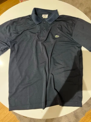 Mörkblå pikétröja från Lacoste - Klassisk mörkblå pikétröja från Lacoste med krage och två knappar framtill. Tröjan har det ikoniska krokodilmärket broderat på bröstet och är tillverkad i bomull för en bekväm känsla. Perfekt till jeans eller shorts.