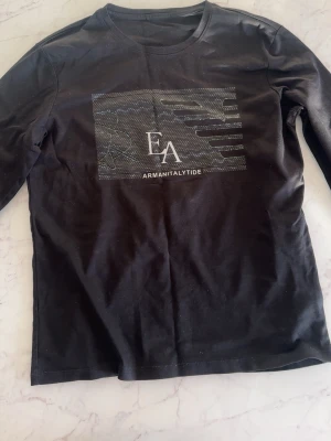 Svart långärmad topp från Armanitalytide - Svart långärmad topp med tryck framtill från Armanitalytide. Toppen har rund halsringning och ett grafiskt mönster med EA-logga på bröstet. Perfekt till jeans eller kjol för en enkel men snygg look.