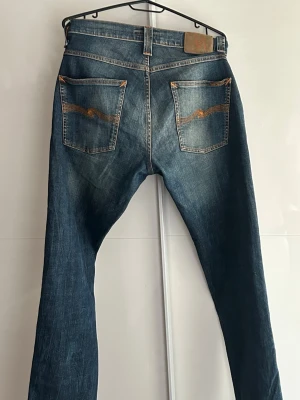 Blå jeans från Nudie Jeans W32 L32 - Säljer ett par klassiska blå jeans från Nudie Jeans med snygg slitning och kontrastsömmar. Jeansen har raka ben, fem fickor och coola broderade detaljer på bakfickorna. Perfekta för en avslappnad och trendig stil.