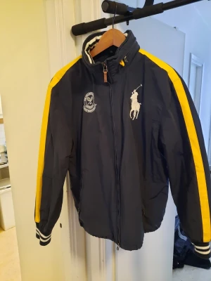 Mörkblå vindjacka Polo Ralph Lauren - Snygg mörkblå vindjacka från Polo Ralph Lauren med gula ränder längs ärmarna och vita detaljer. Jackan har dragkedja, hög krage med randigt foder och broderade loggor på bröstet. Perfekt för dig som vill ha en sportig och stilren look. Storlek Medium