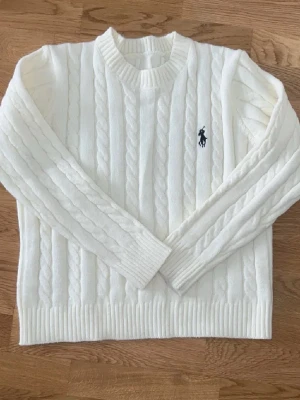 Vit kabelstickad tröja Polo Ralph Lauren - Kabelstickad vit tröja från Polo Ralph Lauren med rund halsringning och svart broderad logga på bröstet. Tröjan har långa ärmar, ribbade muddar och är gjord i ett mjukt material som känns lyxigt och klassiskt. Obs passar inte över 165cm lång kontakta för mer info!!!!