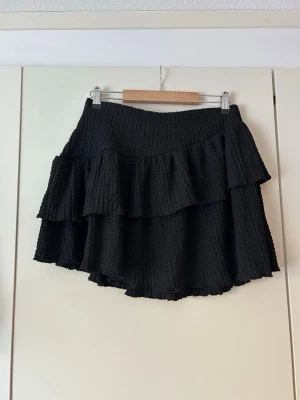 Svart volangkjol med struktur - Säljer en svart kortkjol med dubbla volanger och snygg struktur i tyget. Kjolen har en lätt utsvängd passform och känns både trendig och bekväm. Perfekt att styla med en enkel topp eller stickad tröja.