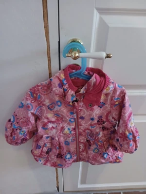 Rosa blommig jacka med huva - Söt rosa jacka med blommigt och paisleymönster, dragkedja framtill och huva. Jackan har puffiga ärmar och elastisk midja för en bekväm passform. Perfekt för att sticka ut med färgglada detaljer och lekfull design.