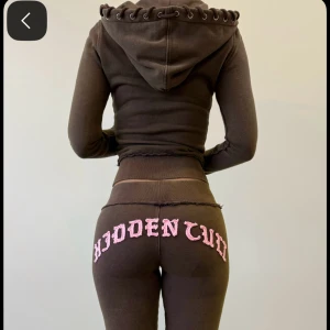 Hidden Cult set - Tvådelat loungewear-set i brun färg från Hidden Cult. Setet består av en croppad hoodie med huva och snörning samt matchande byxor med rosa texten 'HIDDEN CULT' bak på rumpan. Endast använda ett fåtal gånger. Dom är limited edition å går därmed inte längre att köpa. I storlek xxs men passar mer som en xs