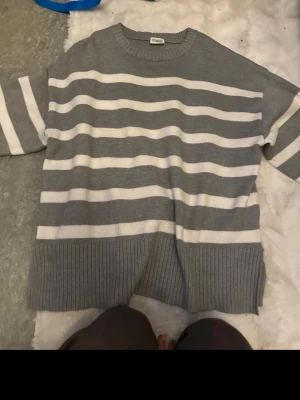 Gråvit randig stickad tröja - Säljer en oversized stickad tröja i grått och vitt med breda horisontella ränder. Tröjan har rund halsringning, ribbstickade muddar och en avslappnad passform. Perfekt för en chill och stilren look.