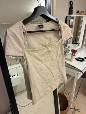 Beige croppad topp från Ginatricot - Trendig beige croppad topp från Ginatricot med korta ärmar och fyrkantig ringning. Toppen har dekorativa knytband på axlarna och är tillverkad i mjuk bomull. Perfekt för en avslappnad och stilren look.