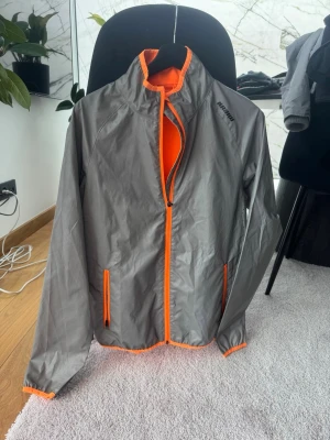 Grå och orange vindjacka från Karhu - Snygg vindjacka i grått med starka orange detaljer från Karhu. Jackan har hel dragkedja, två fickor med orange dragkedjor och en cool kontrastfärgad baksida. Lätt material som passar perfekt för utomhusaktiviteter. Sportig look med hög krage och elastiska muddar.