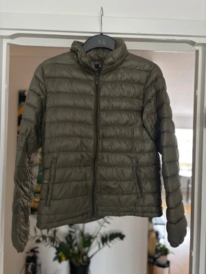 Olivgrön pufferjacka från H&M - Snygg olivgrön pufferjacka från H&M med quiltad design och dragkedja framtill. Jackan har hög krage och två sidofickor med dragkedja. Perfekt för kalla dagar och enkel att matcha med olika outfits.