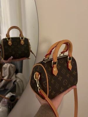 Louis Vuitton Speedy Nano Monogram - Säljer en Louis Vuitton Speedy Nano handväska i klassiskt brunt monogrammönster med beige och röda detaljer. Väskan har guldfärgade metalldetaljer, två handtag och avtagbar axelrem. Tillverkad i canvas med läderdetaljer. Perfekt för dig som gillar lyxig och tidlös design.