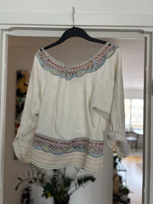 Vit boho-blus med brodyr och paljetter - Vit bohemisk blus med färgglad brodyr i lila, blått och beige längs halsringning och nederkant. Blusen har lång ärm och en lös passform. Dekorativ detalj med guldpaljetter framtill. Perfekt för en avslappnad och trendig stil.